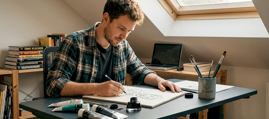 Ein Künstler sitzt an seinem Schreibtisch im Atelier und bringt eine Karikatur aufs Papier.